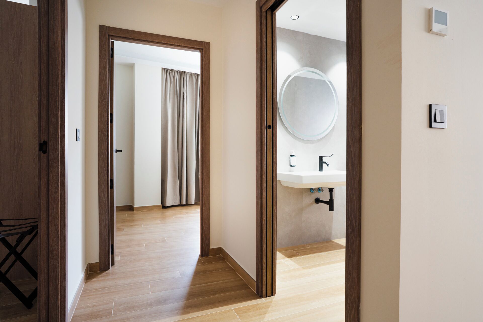 A hotel room with a wooden floor, a bathroom with a round mirror and sink, and a door leading to another room.