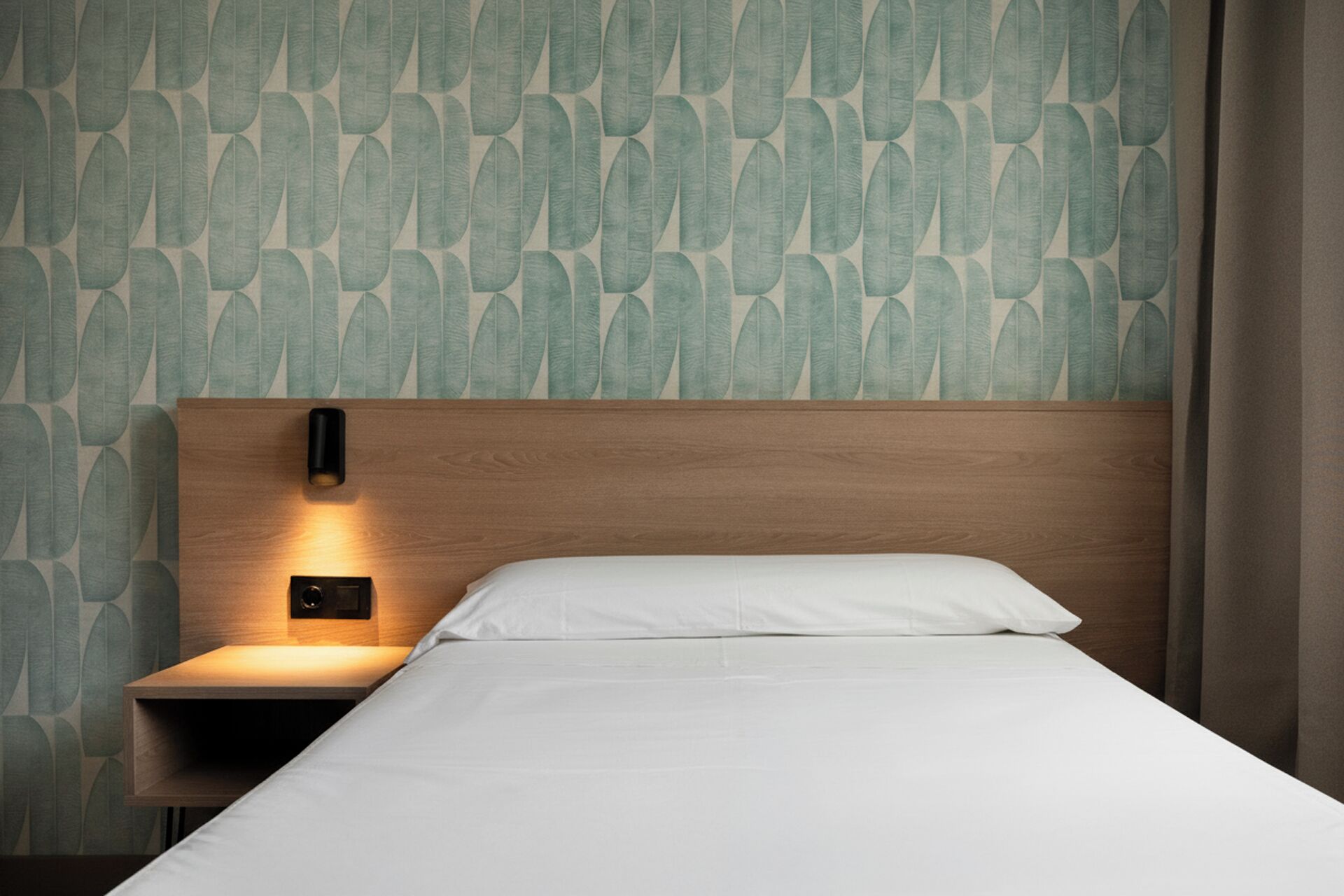 Cama bem arrumada com cabeceira de madeira e mesa de cabeceira com luminária fixada na parede.