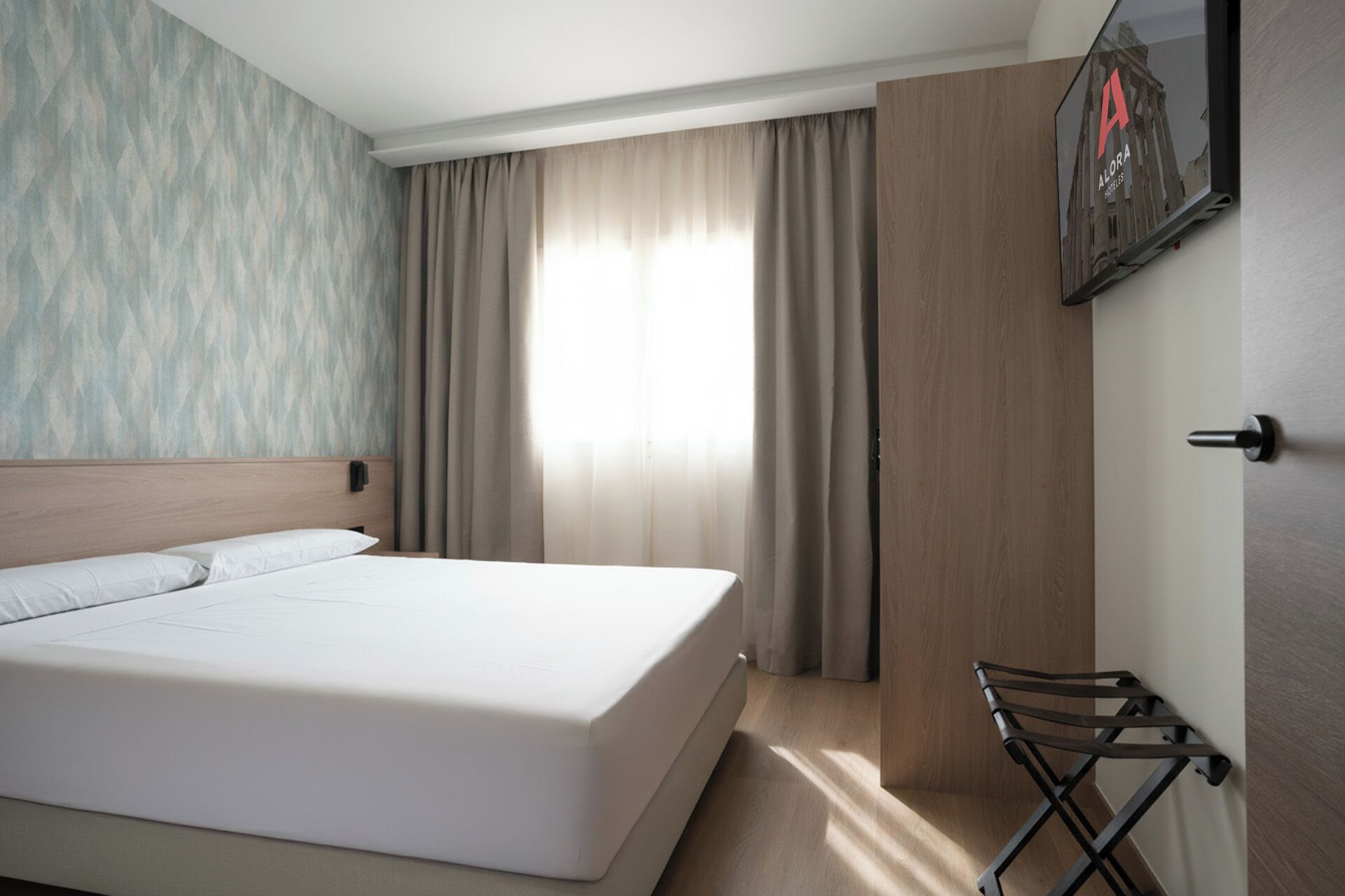 Una habitación de hotel con cama, cortinas, televisor montado en la pared y una puerta de madera.