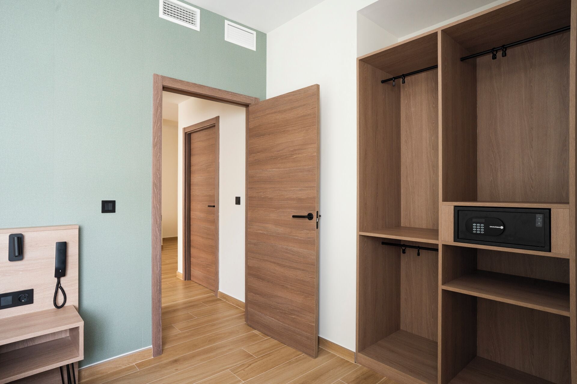 A room with a wooden wardrobe, a desk with a telephone, and a door leading to another room.