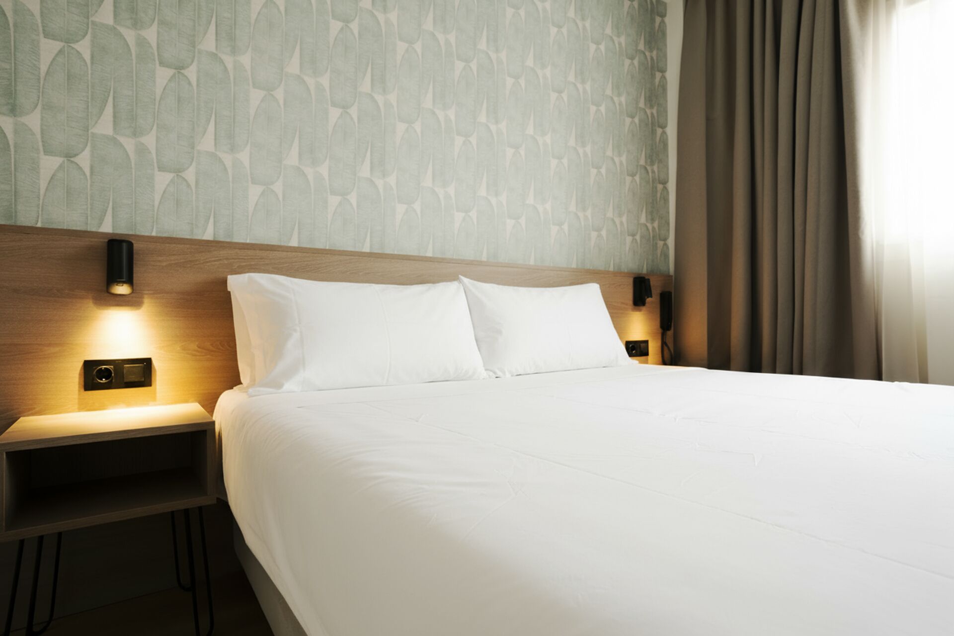 A hotel room with a large bed, two bedside tables with lamps, a wooden headboard, and a patterned wallpaper.