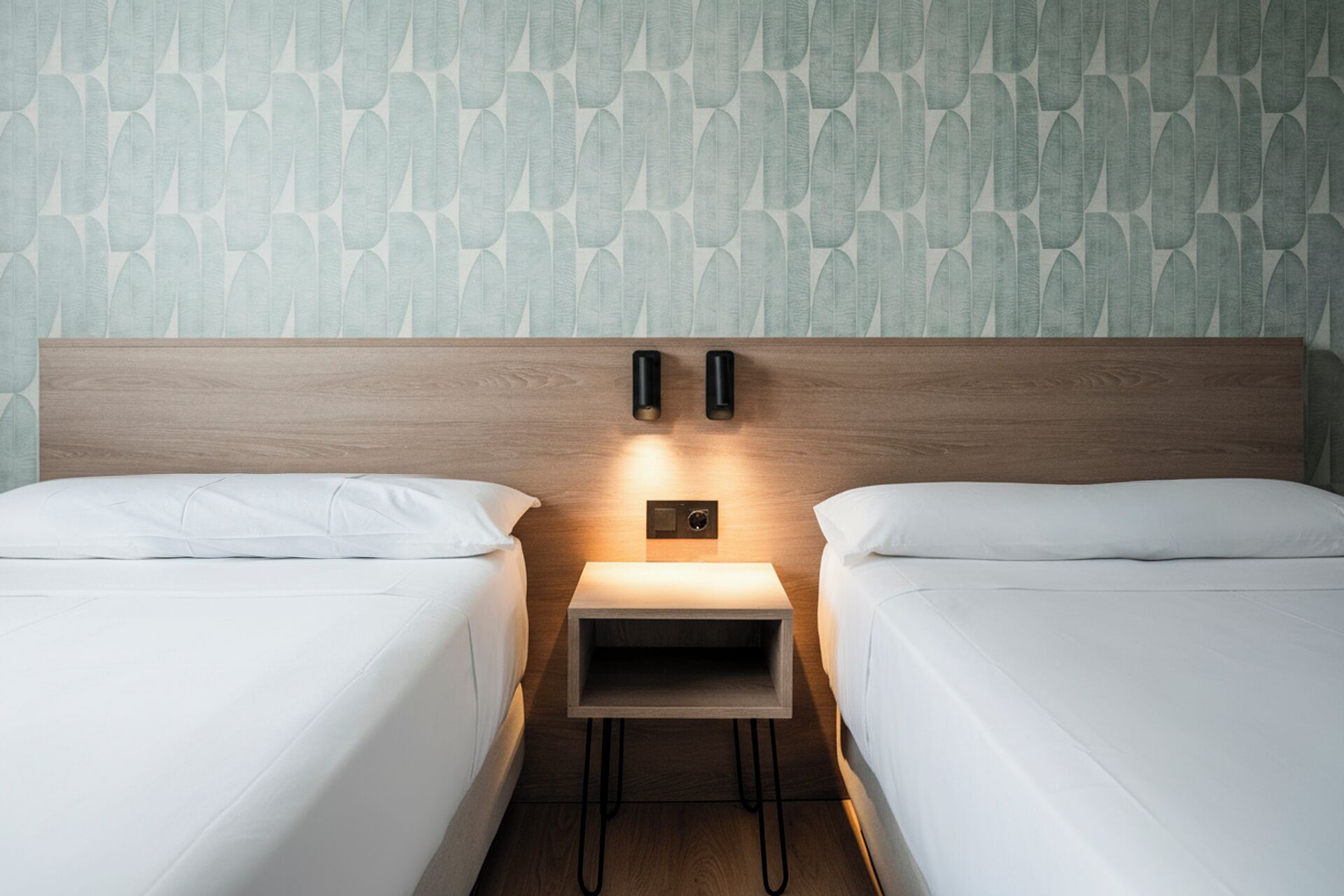 A hotel room with two single beds, a wooden headboard, a nightstand with a lamp, and a patterned wall.