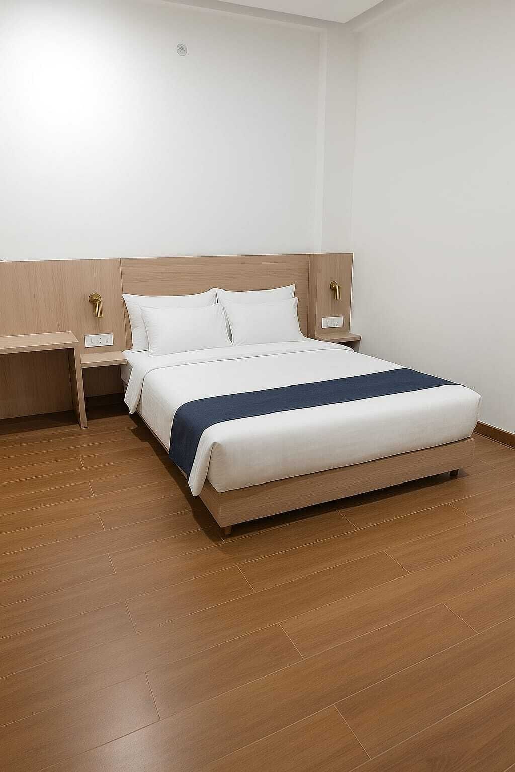 Habitación de hotel con piso de madera, cama con ropa blanca y un detalle azul, dos mesitas de noche con lámparas y una pared blanca lisa.