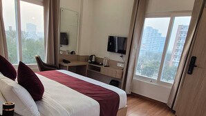 A modern hotel room with a large bed, wooden flooring, a ceiling fan, a desk, a chair, a TV, and large windows offering a city view. - Darpan Nikunj Rubystone Express (Vrindavan)