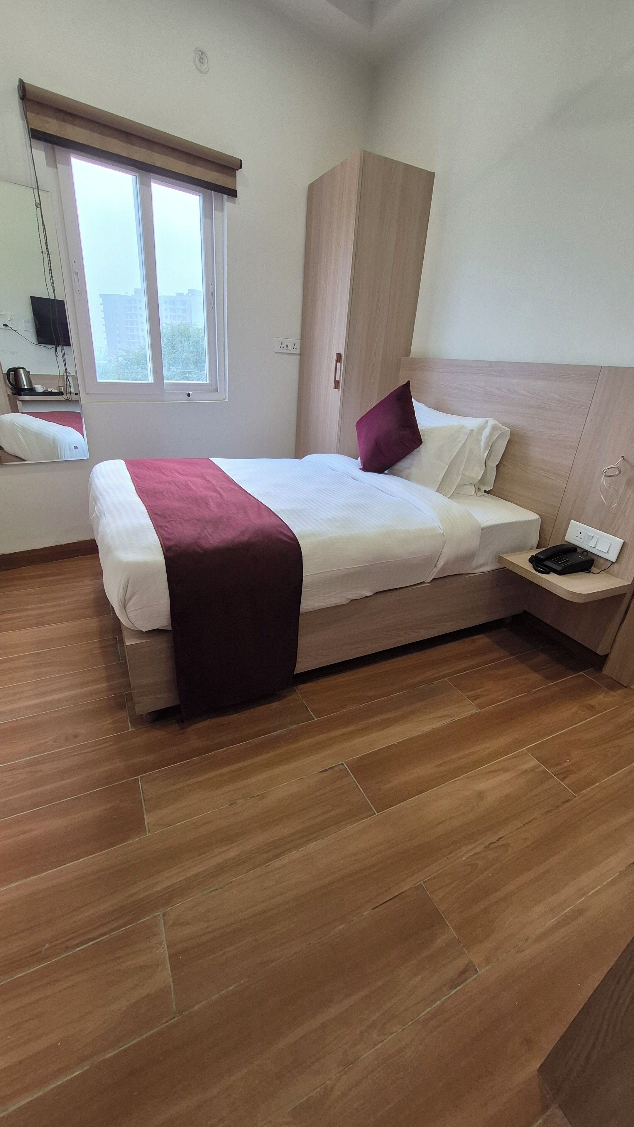 Een moderne hotelkamer met een groot bed, een houten garderobe, een nachtkastje met telefoon en een raam met zonwering.
