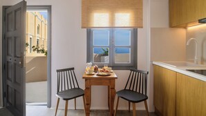 Een kleine keuken met een houten tafel gedekt voor het ontbijt, een raam met zonwering, en uitzicht op een gebouw.