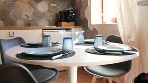 Ein Essbereich mit einem runden Tisch, gedeckt für vier Personen, umgeben von Stühlen. Der Tisch ist weiß, die Stühle schwarz. Im Hintergrund steht eine Kaffeemaschine auf der Arbeitsplatte.