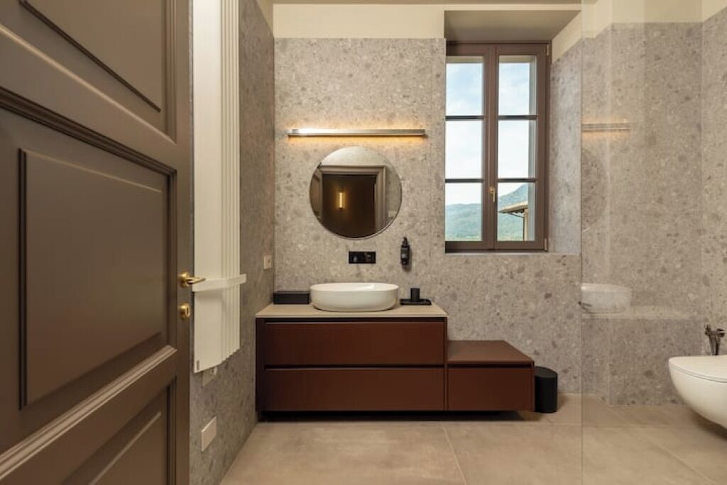A modern bathroom with a marble wall, a round mirror, a built-in sink, and a window.