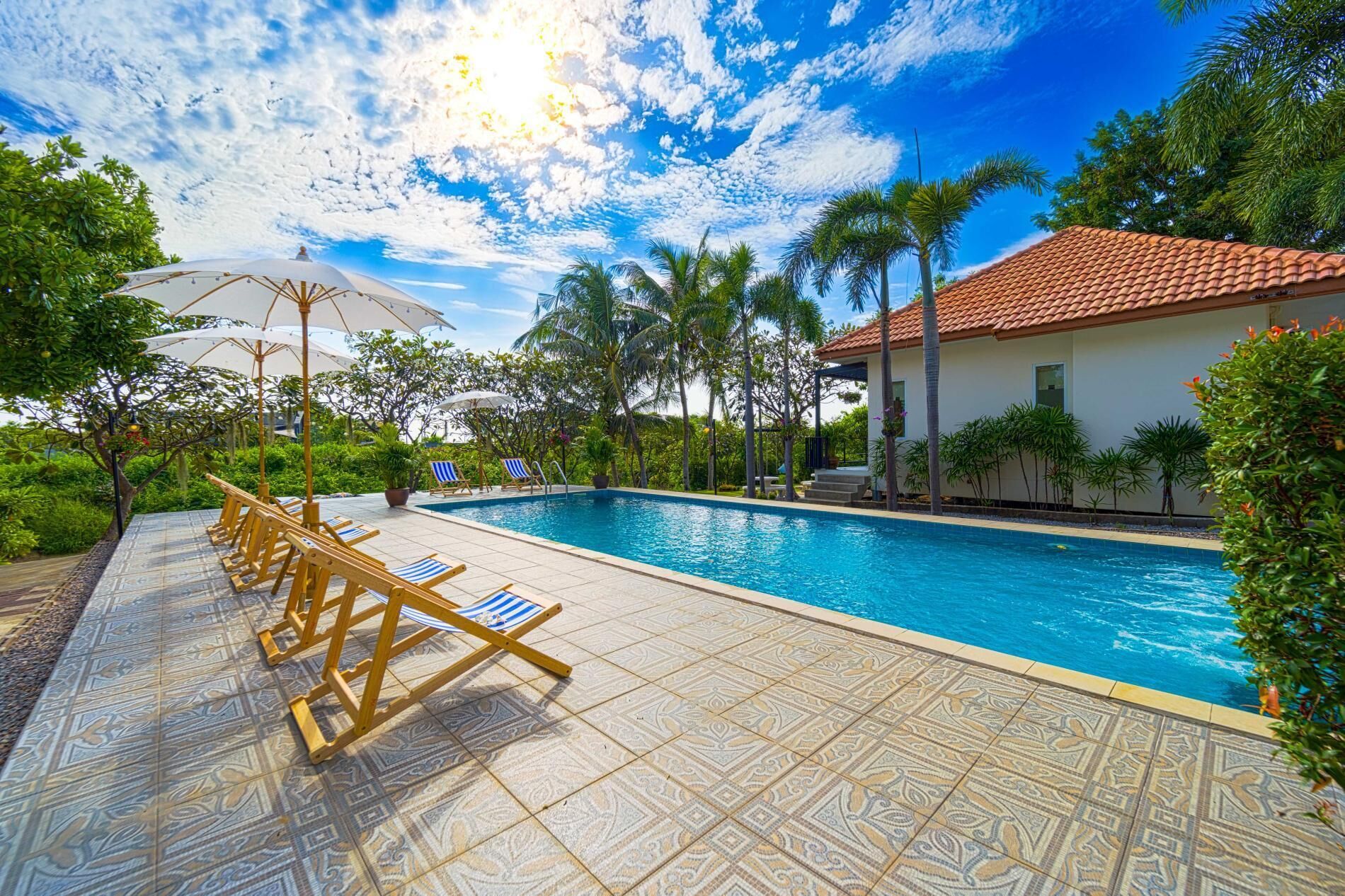 A swimming pool with lounge chairs and umbrellas, surrounded by palm trees and a building with a tiled roof.