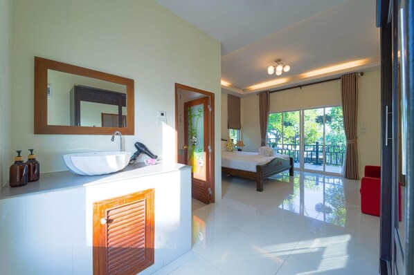 A modern hotel room with a large bed, a bathroom with a sink and mirror, and a balcony with a view of greenery.