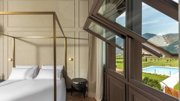 A bedroom with a four-poster bed, a small round table, and a view of a swimming pool and mountains through large windows. - Suite..... (Iseo)