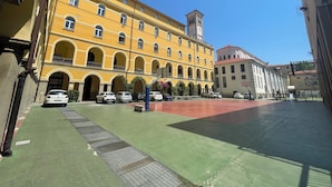 A courtyard with a yellow building featuring arched windows and balconies, a clock tower, and a basketball court.
