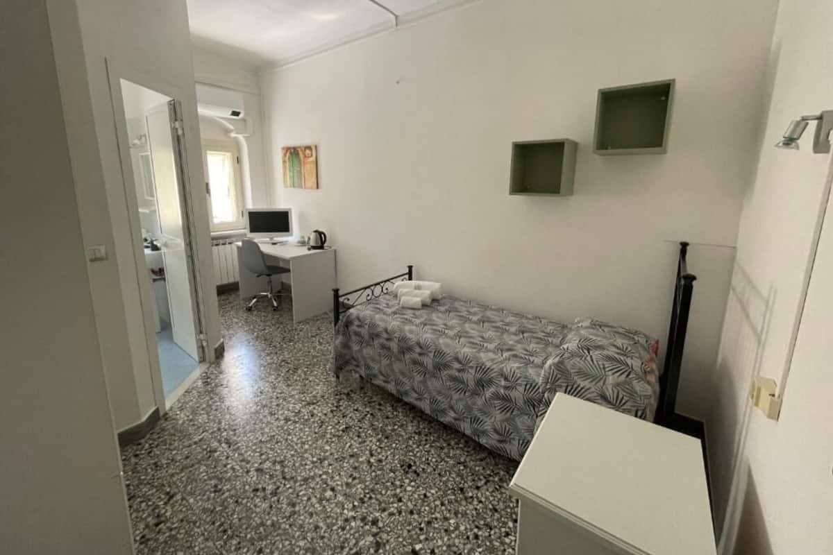 A small, compact room with a bed, a desk, and a chair. There is a small window and a door leading to another room.