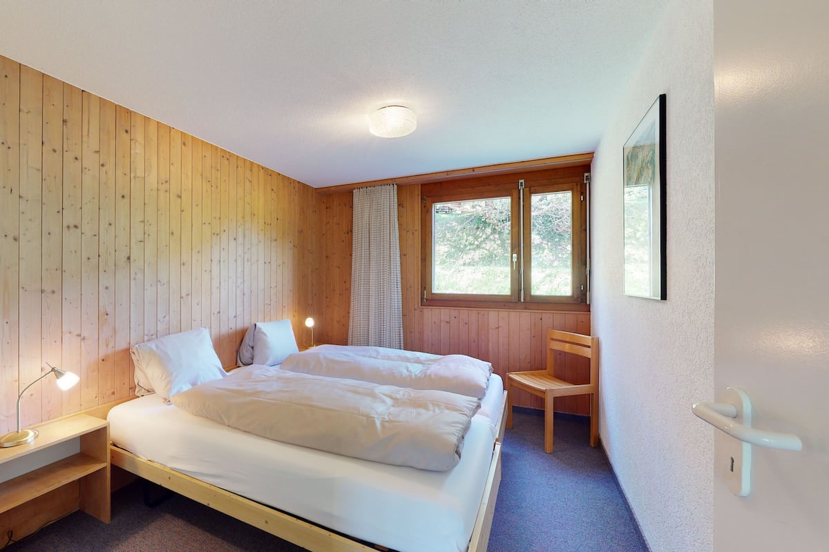 Ein Schlafzimmer mit einem Holzbett, einem Nachttisch mit Lampe, einem Stuhl und einem Fenster mit Vorhängen.