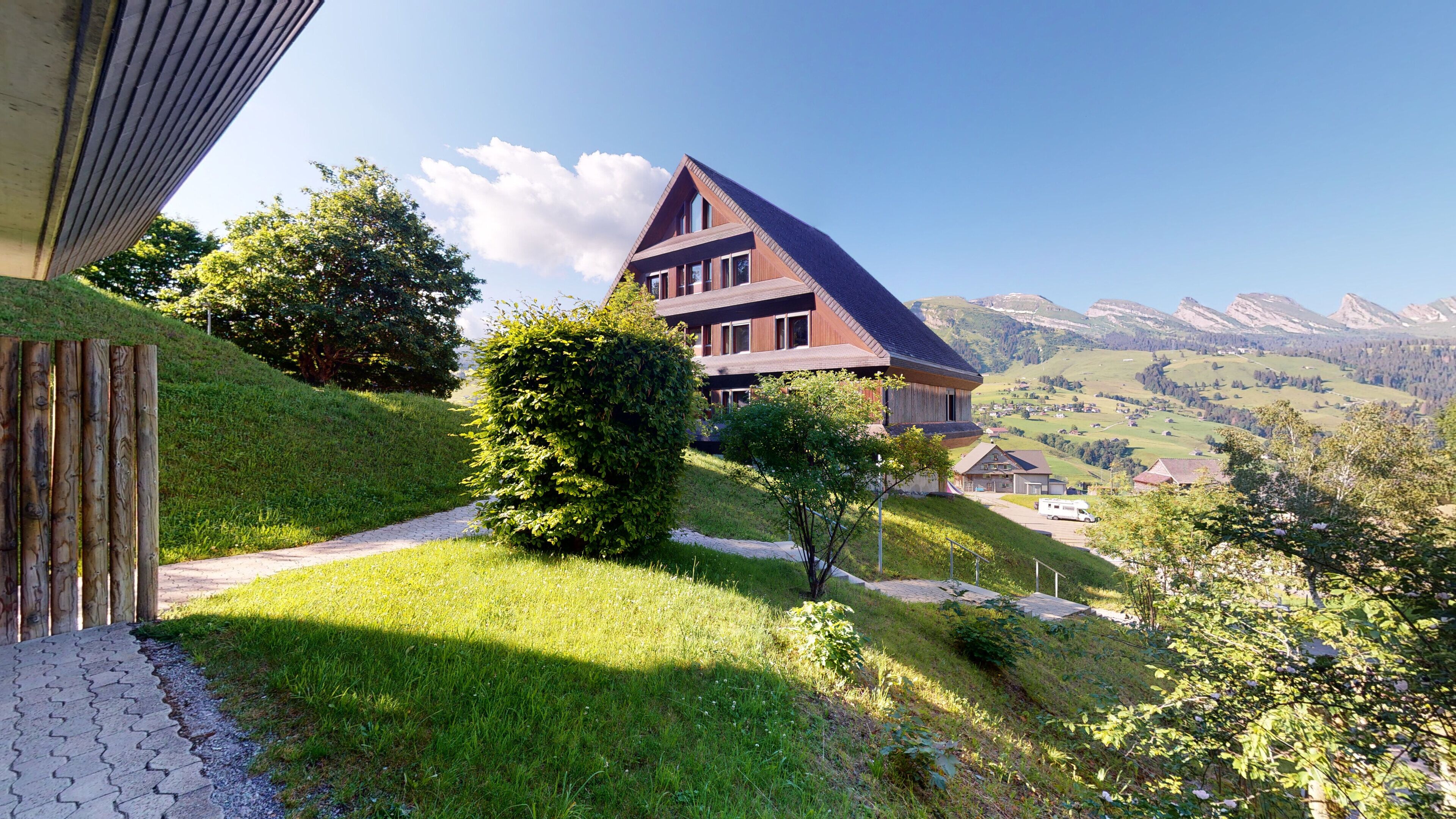 Une maison moderne, dotée d’un toit en pente, entourée d’une végétation luxuriante et offrant une vue dégagée sur les montagnes lointaines.