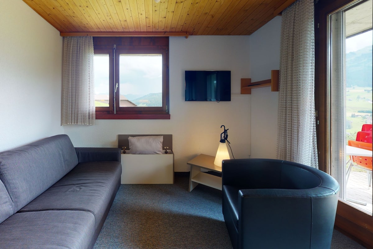 Ein Zimmer mit grauem Sofa, einem kleinen Nachttisch mit Lampe, einem an der Wand montierten Fernseher und einem Fenster mit Vorhängen.