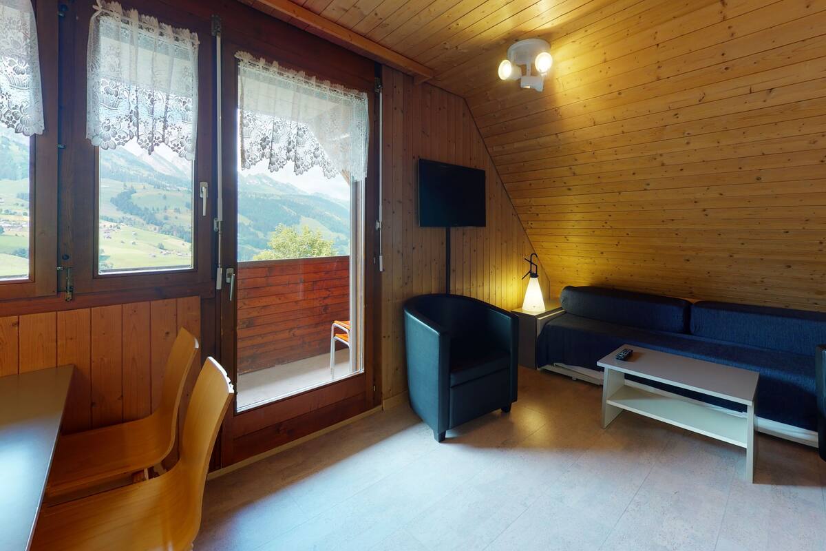 Ein gemütliches Zimmer mit Holzbalkendecke, einer Couch, einem Tisch und einem Stuhl. Ein Fernseher ist an der Wand befestigt, und es gibt ein Fenster mit Spitzenvorhängen.