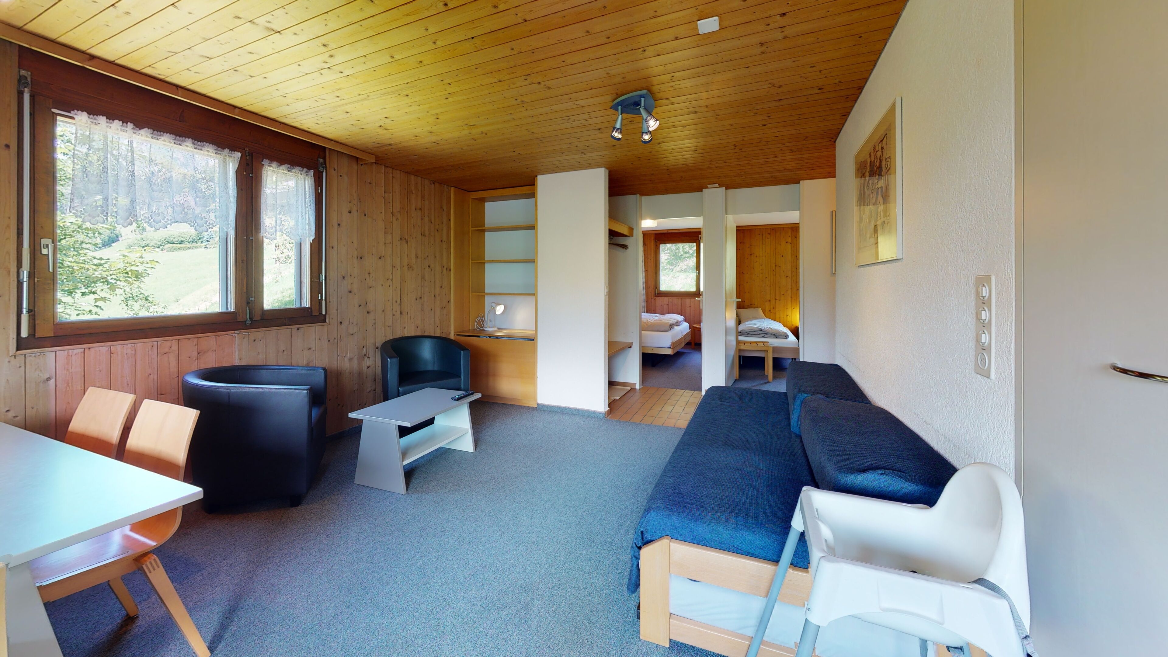 A cozy room with a wooden ceiling, a sofa, a table, and a chair. There is a bed in the background and a window with lace curtains.