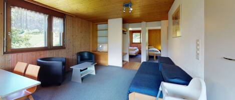 A cozy room with a wooden ceiling, a sofa, a table, and a chair. There is a bed in the background and a window with lace curtains.