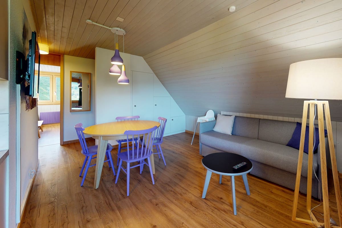 Ein gemütliches Zimmer mit Holzboden, einem runden Tisch mit Stühlen, einer Couch und einer Stehlampe.