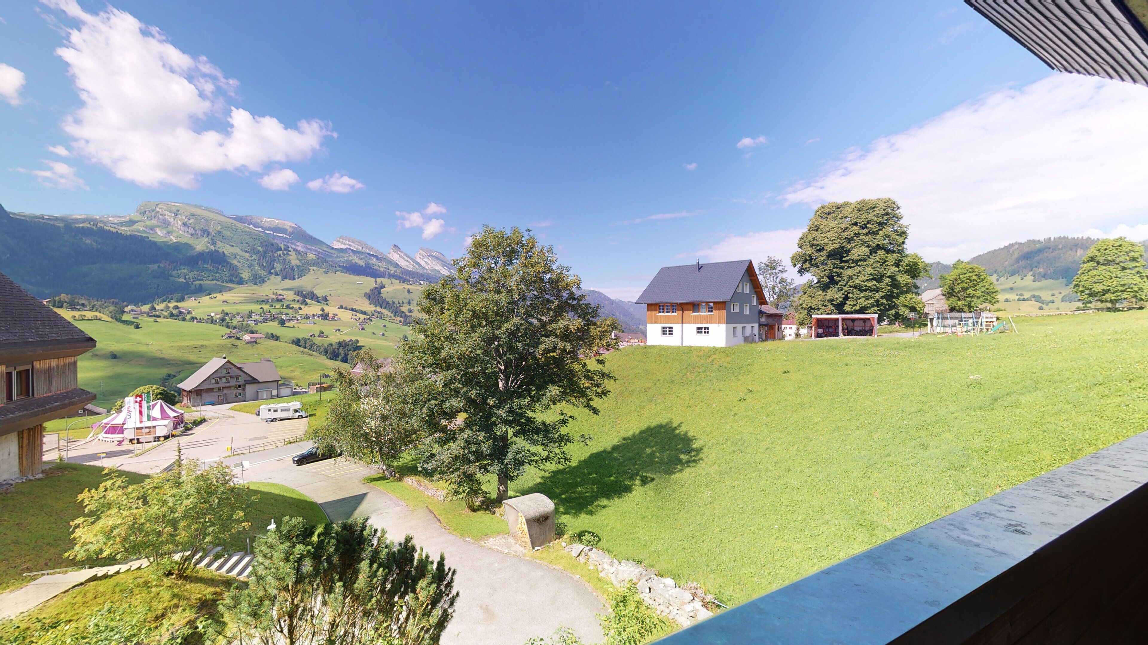Ländliche Landschaft mit einem Haus, einem Baum und einem Gebirgszug im Hintergrund.