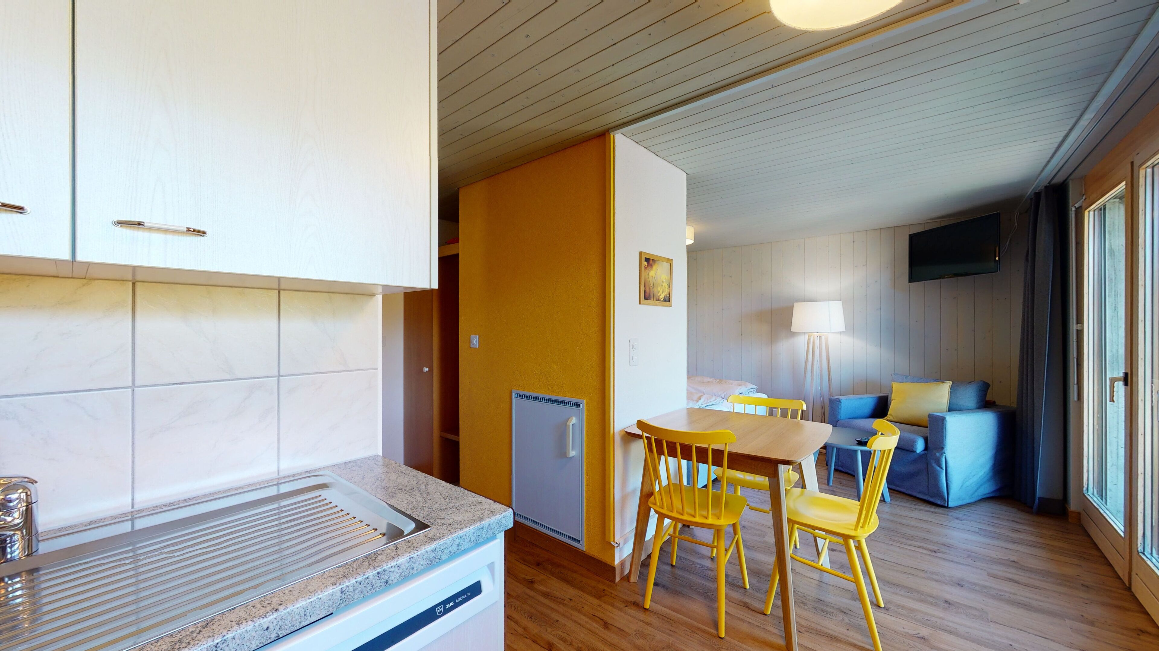 Une cuisine compacte dotée d’un four à micro-ondes encastré et d’un petit réfrigérateur. On y trouve un coin repas avec une table en bois et des chaises jaunes. La pièce est chaleureuse, avec un canapé bleu et une lampe.
