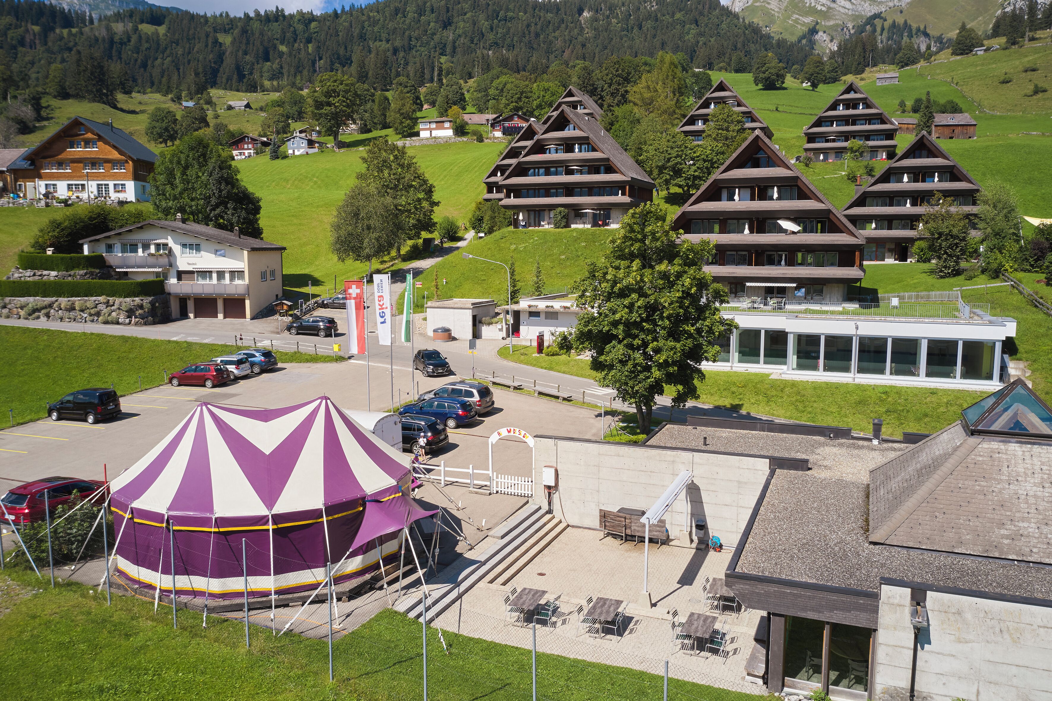 A mountainous village with traditional chalet-style buildings, a parking area, and a large tent structure.