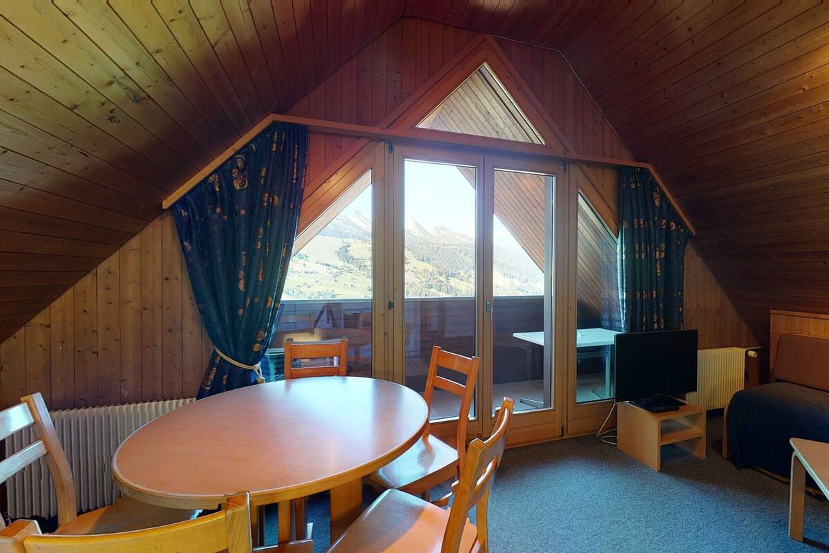 Ein gemütliches Dachzimmer mit Holzvertäfelung, einem runden Tisch, Stühlen, einer Couch und einem Fernseher.