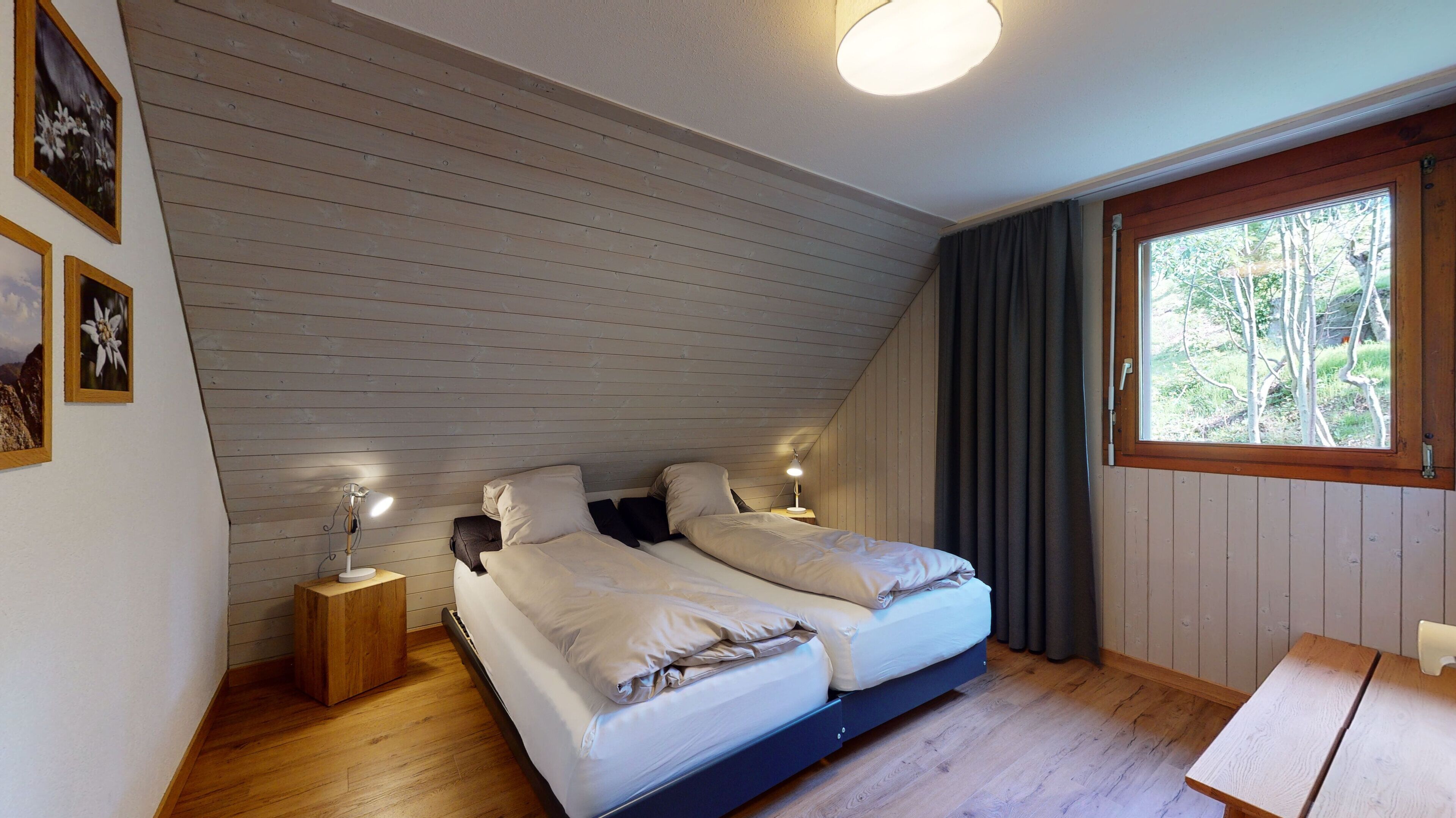 A bedroom with two beds, wooden walls, a window with curtains, and a bedside table with a lamp.