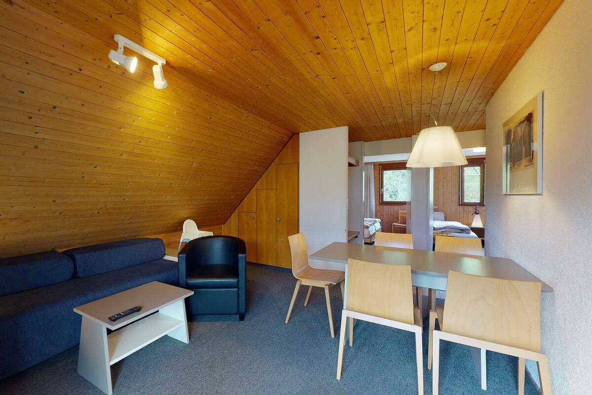 Ein gemütlicher, gut beleuchteter Raum mit Holzbalkendecke, einer Couch, einem Esstisch mit Stühlen und einem kleinen Tisch für den Fernseher.