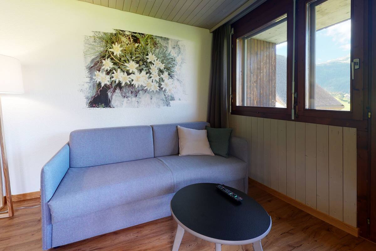 Ein Zimmer mit grauem Sofa, einem kleinen runden Tisch mit Fernbedienung, einer Stehlampe und einem Fenster mit Bergblick.