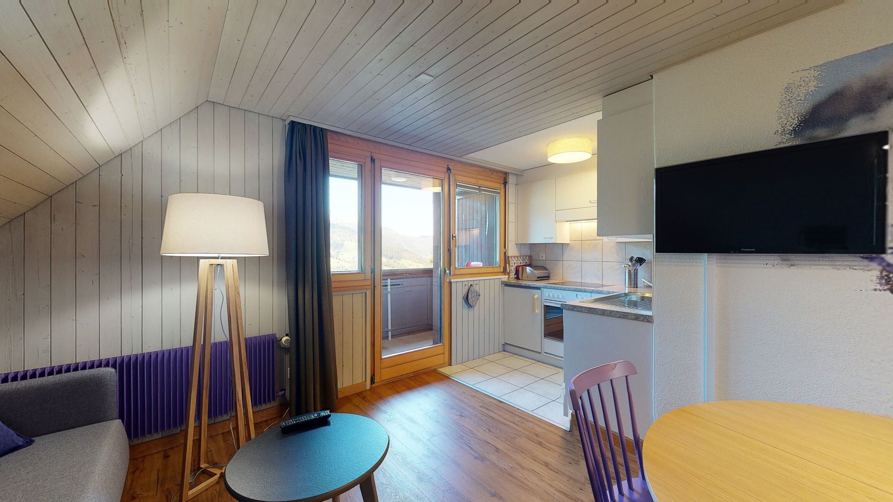 A cozy room with a wooden ceiling, a sofa, a small table, a lamp, a TV, and a kitchen area with a sink and stove.