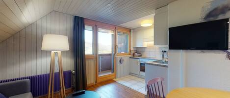 A cozy room with a wooden ceiling, a sofa, a small table, a lamp, a TV, and a kitchen area with a sink and stove.