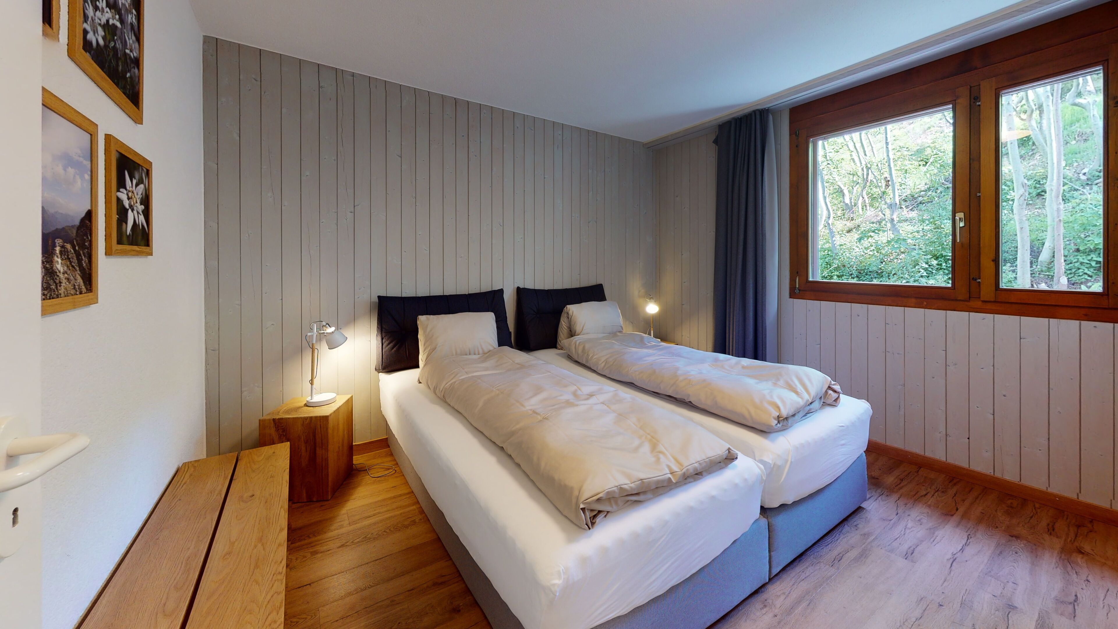 Un dormitorio con dos camas, suelo de madera, una mesita de noche con lámpara y una ventana con vistas a árboles.