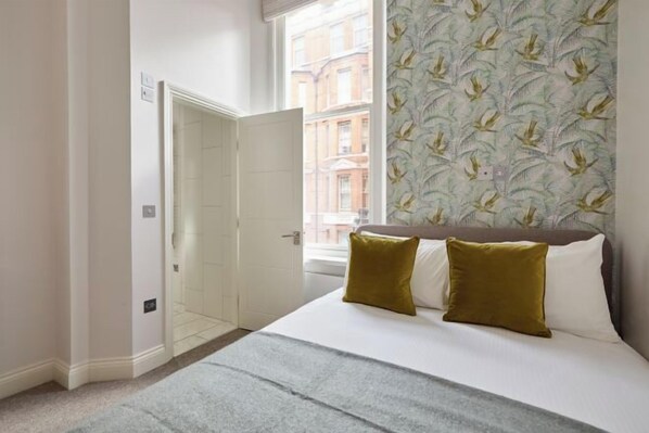 A bedroom with a bed, two pillows, a window with a view of buildings, and a door. - 2VH Virginia House, 31 Bloomsbury Way by City Living London (London)