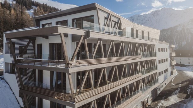Ein modernes, mehrstöckiges Gebäude mit Holzfassade und großen Fenstern, vor einer verschneiten Berglandschaft.