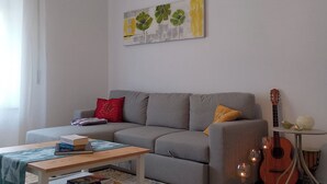 Een woonkamer met een grijze bank, een bijzettafeltje met boeken, een gitaar en een kunstwerk aan de muur.