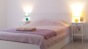 Een slaapkamer met een wit bed, twee nachtkastjes met lampen, een hanglamp en een vloerkleed met patroon.