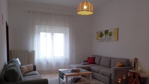 Een woonkamer met een grijze bank, een witte salontafel, en een grijze zithoek. Er staat een kleine tafel met kaarsen en een gitaar.