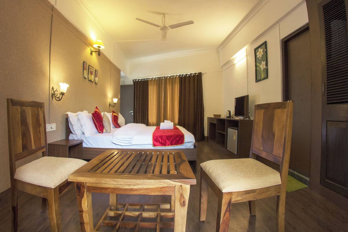 A hotel room with a bed, two wooden chairs, a small table, a desk with a chair, a ceiling fan, and a television.