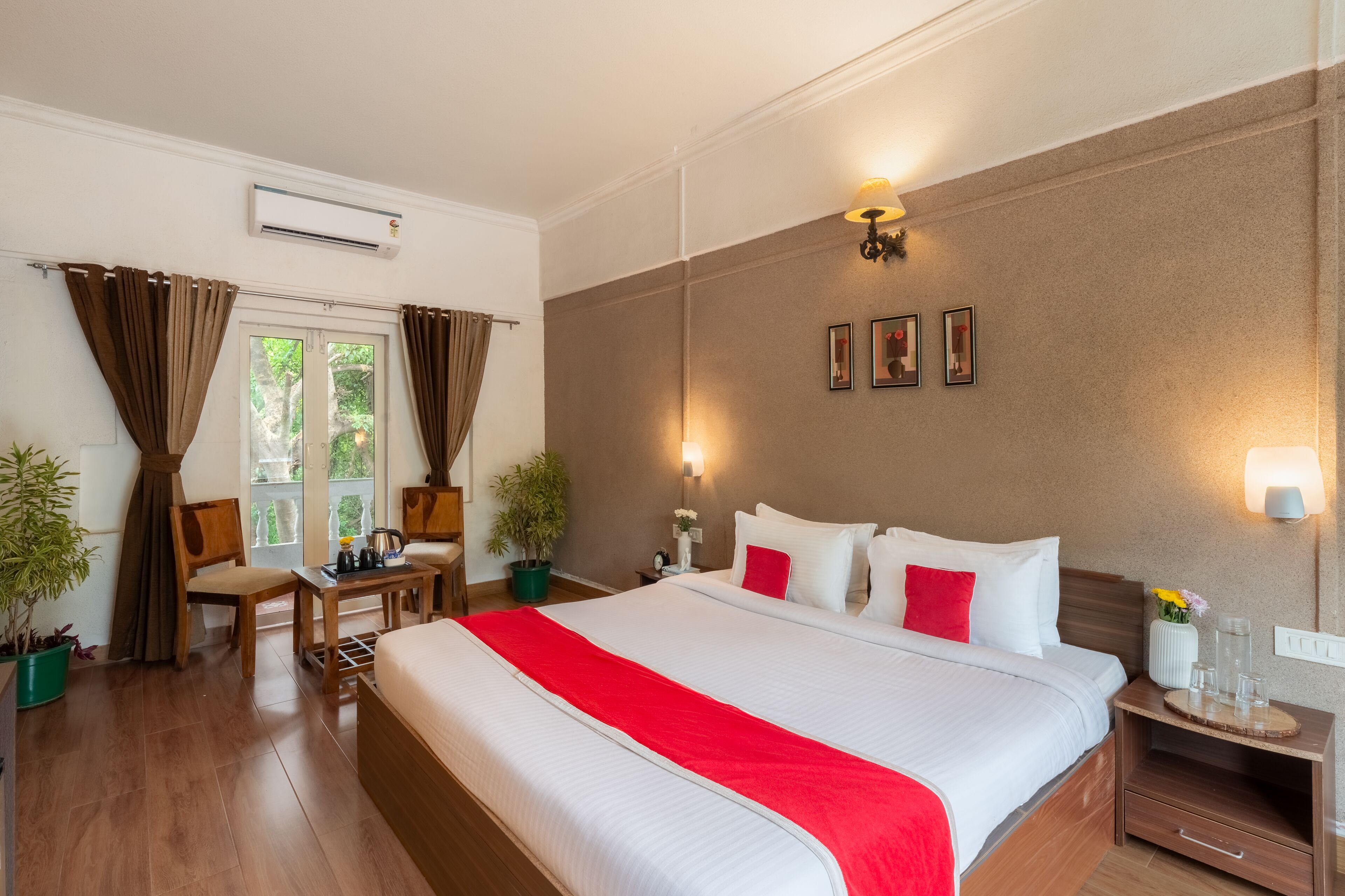 A hotel room with a large bed, a desk, two chairs, a potted plant, a nightstand, and a wall-mounted air conditioner.