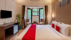 A hotel room with a bed, a TV, a desk, a small fridge, a potted plant, a window with curtains, and a wall-mounted air conditioner. - Velvet County Resort (Lonavala)
