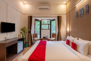 Une chambre d’hôtel équipée d’un lit, d’une télévision, d’un bureau, d’un petit réfrigérateur, d’une plante en pot, d’une fenêtre avec des rideaux et d’un climatiseur fixé au mur.