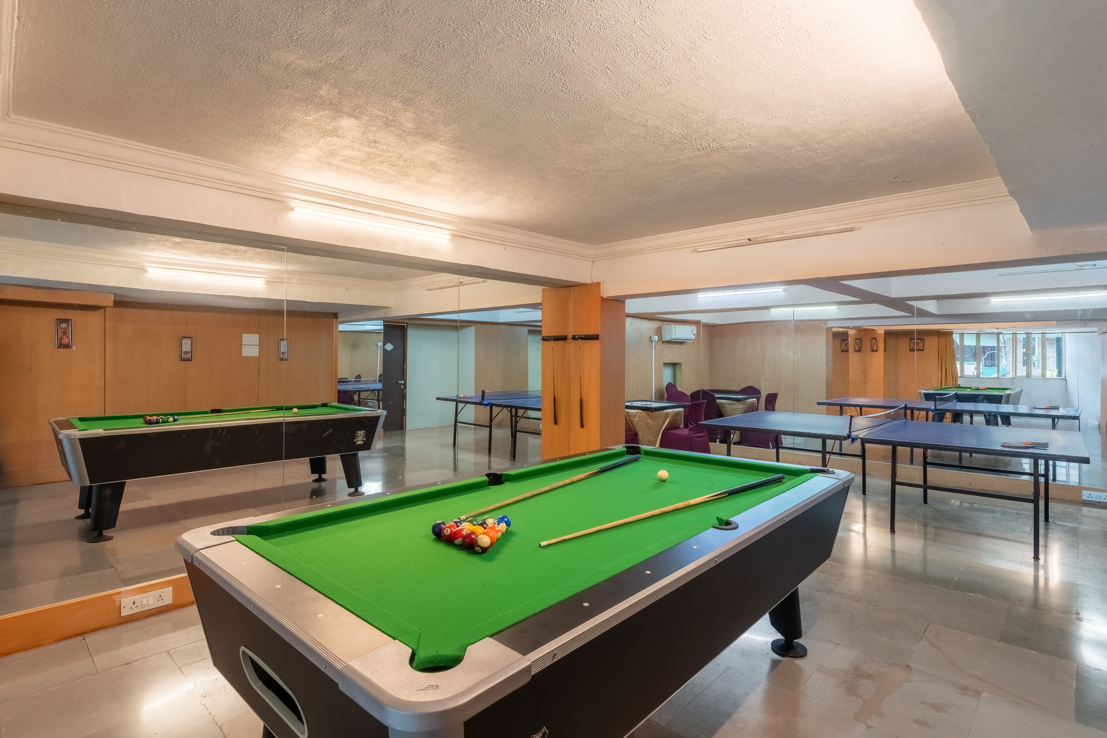 Una mesa de billar con bolas, taco y superficie de fieltro verde. Hay dos mesas de billar, una mesa de ping pong y una zona de estar con sillas.