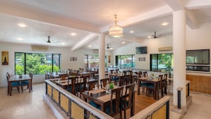 A spacious dining area with multiple tables and chairs, ceiling fans, and large windows offering views of greenery. - Velvet County Resort (Lonavala)