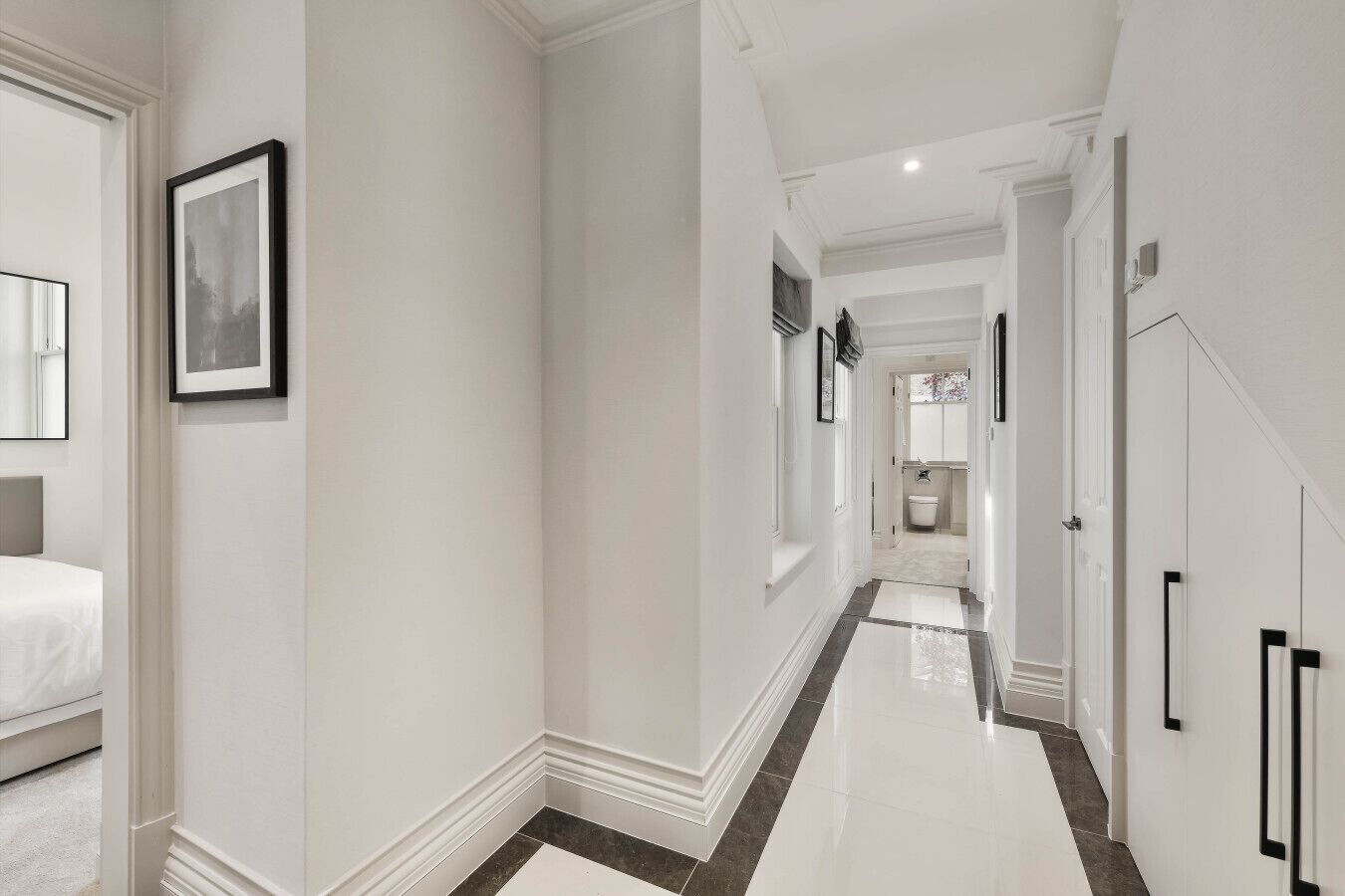 A hallway with a bed on the left, framed pictures on the walls, and a bathroom visible in the distance.