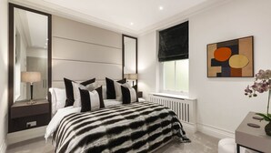A modern bedroom with a large bed, a bedside table, a mirror, a window with a view, and a piece of abstract art on the wall. - Kensington Apartment | Hyde Park, Portobello, Royal Albert Hall (London)