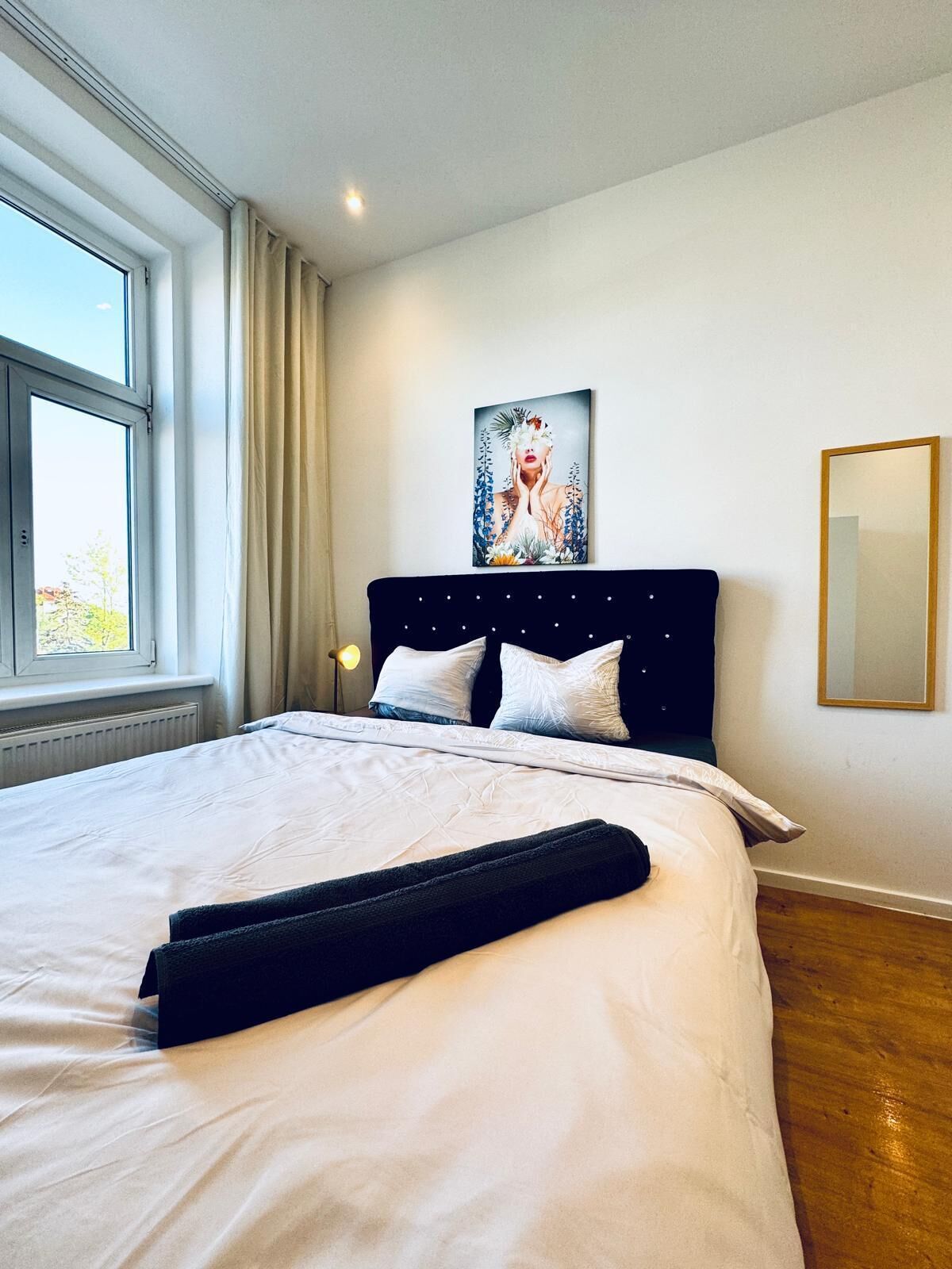 A neatly made bed with white linens, a dark headboard, and a large window with curtains.