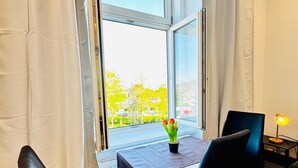 Een kamer met een tafel en twee stoelen, een raam met gordijnen, en een bijzettafeltje met een lamp.