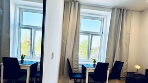 Een moderne, goed verlichte kamer met twee eettafels, zwarte stoelen en een houten bank. Grote ramen met gordijnen laten natuurlijk licht en uitzicht op bomen binnen.